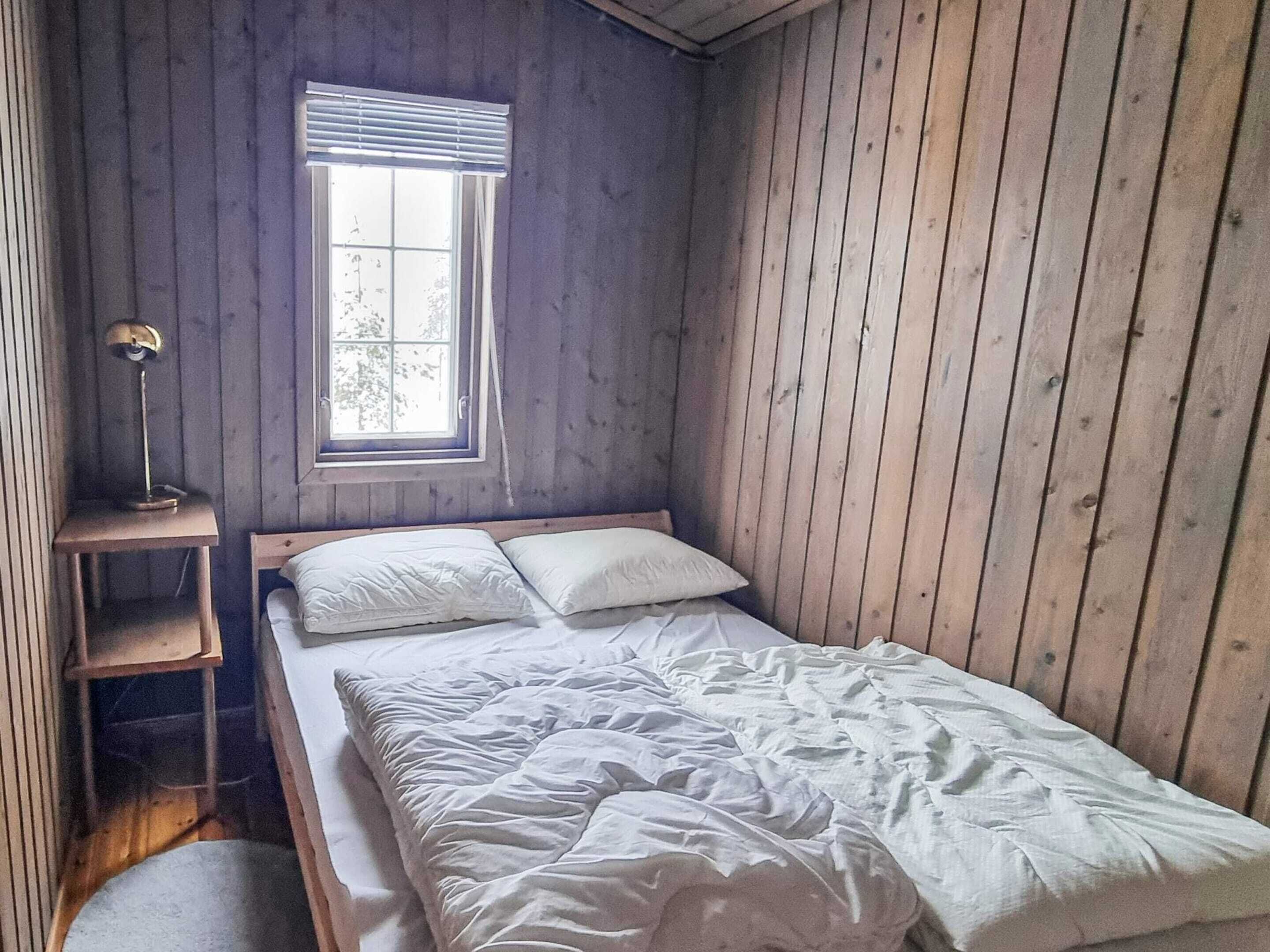 A small, single-bed room with wooden walls, a window with blinds, a bedside table with a lamp, and a small shelf.