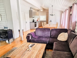 Un salón acogedor con un sofá morado, una mesa de centro de madera y un calentador negro. Se ve una cocina con armarios blancos y una zona de comedor con una mesa y sillas de madera al fondo.