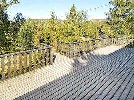 Una terraza de madera con barandilla, rodeada de árboles y un paisaje montañoso al fondo.