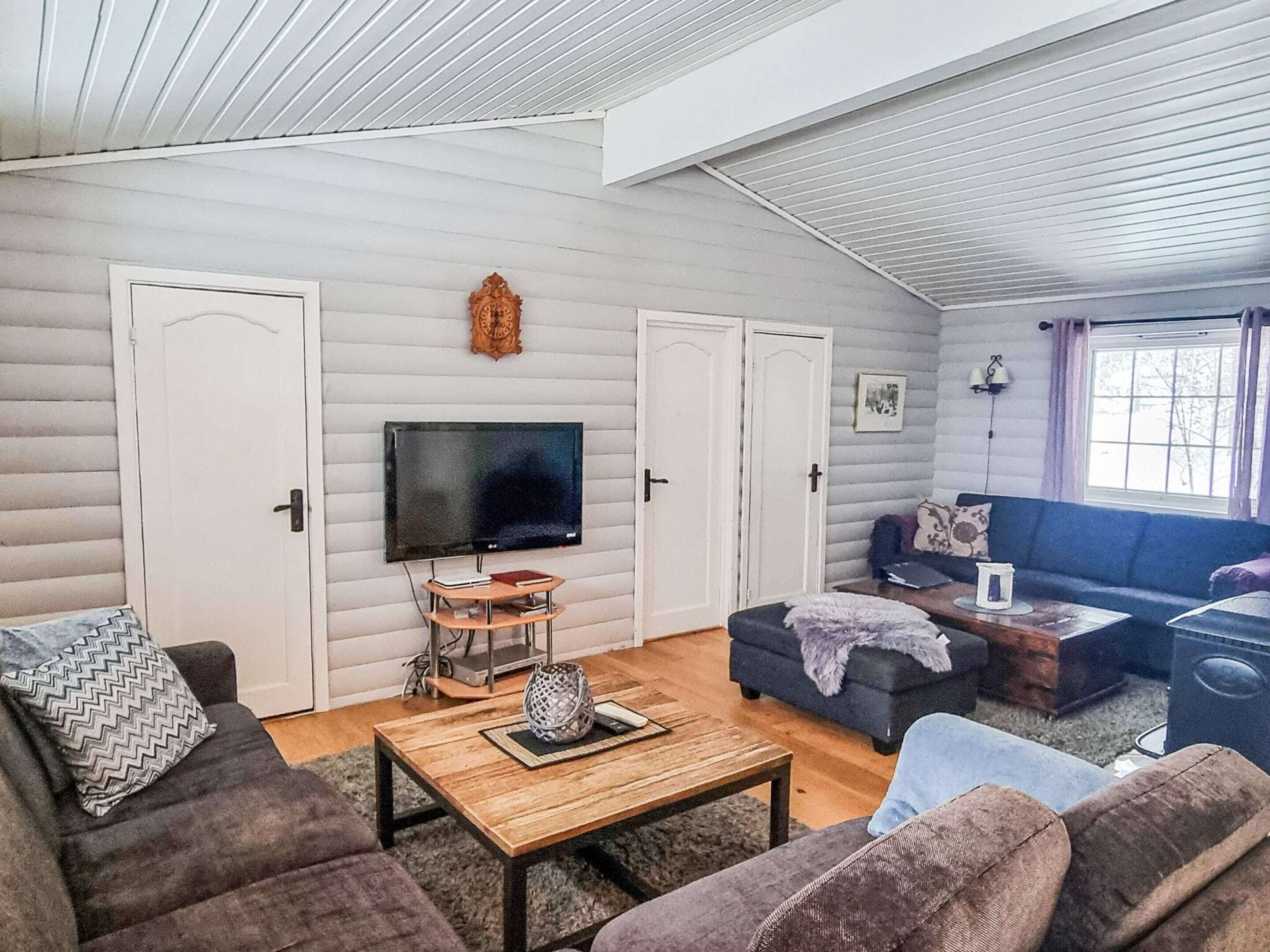 A cozy living room with a flat-screen TV, a wooden coffee table, and a sofa. There are two doors and a window with curtains.
