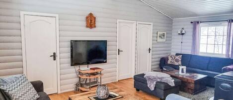 A cozy living room with a flat-screen TV, a wooden coffee table, and a sofa. There are two doors and a window with curtains.