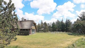 A rustic cabin with a thatched roof, surrounded by a grassy field and dense trees.
