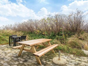 Uma mesa de piquenique de madeira ao ar livre, com bancos, rodeada por uma paisagem natural com arbustos e árvores.