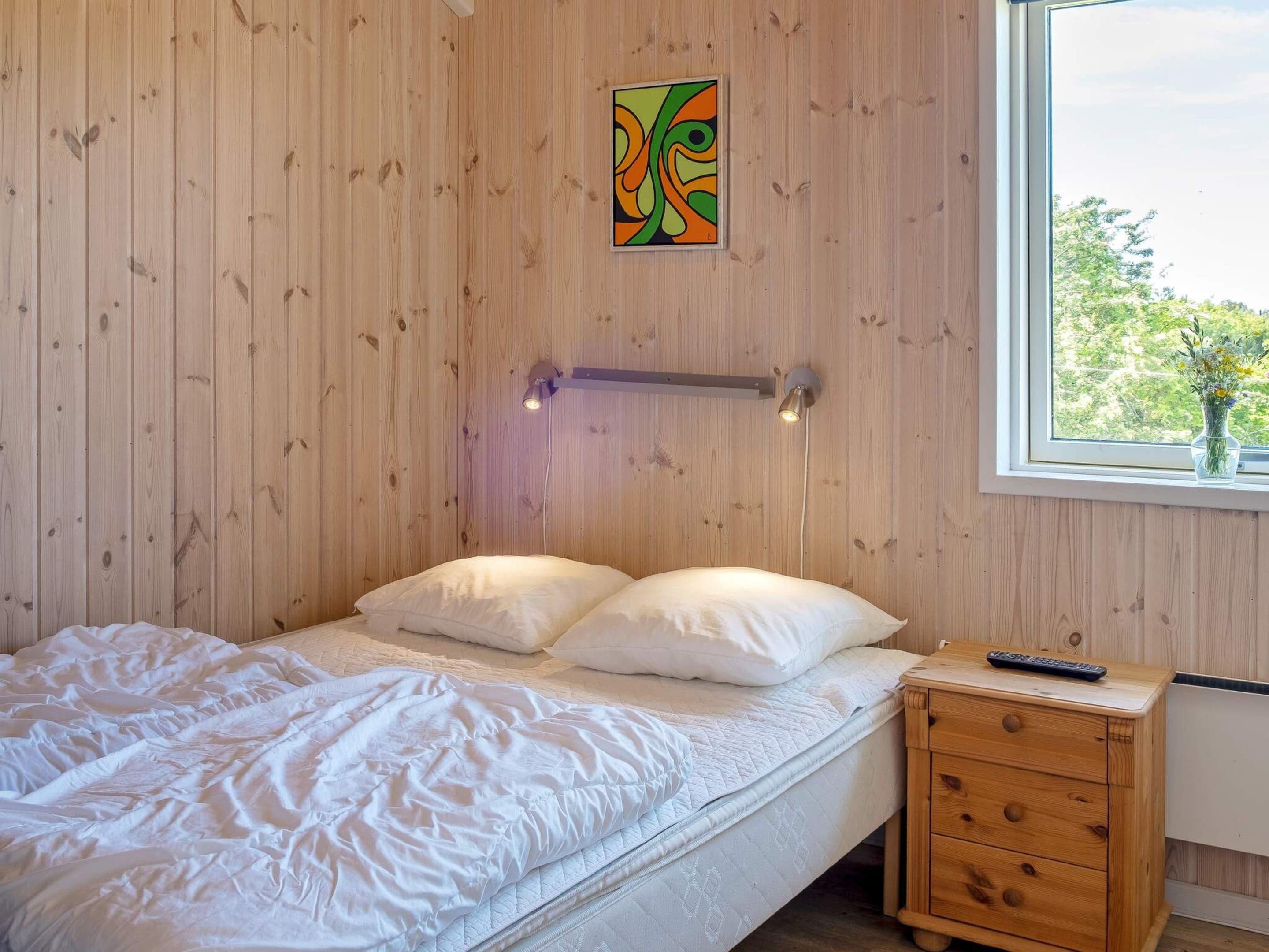 A bedroom with a bed, a nightstand, a wall-mounted lamp, and a window with a view of greenery.