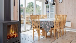 A dining area with a wood-burning stove, wooden chairs, a table with a floral tablecloth, and a view through glass doors.