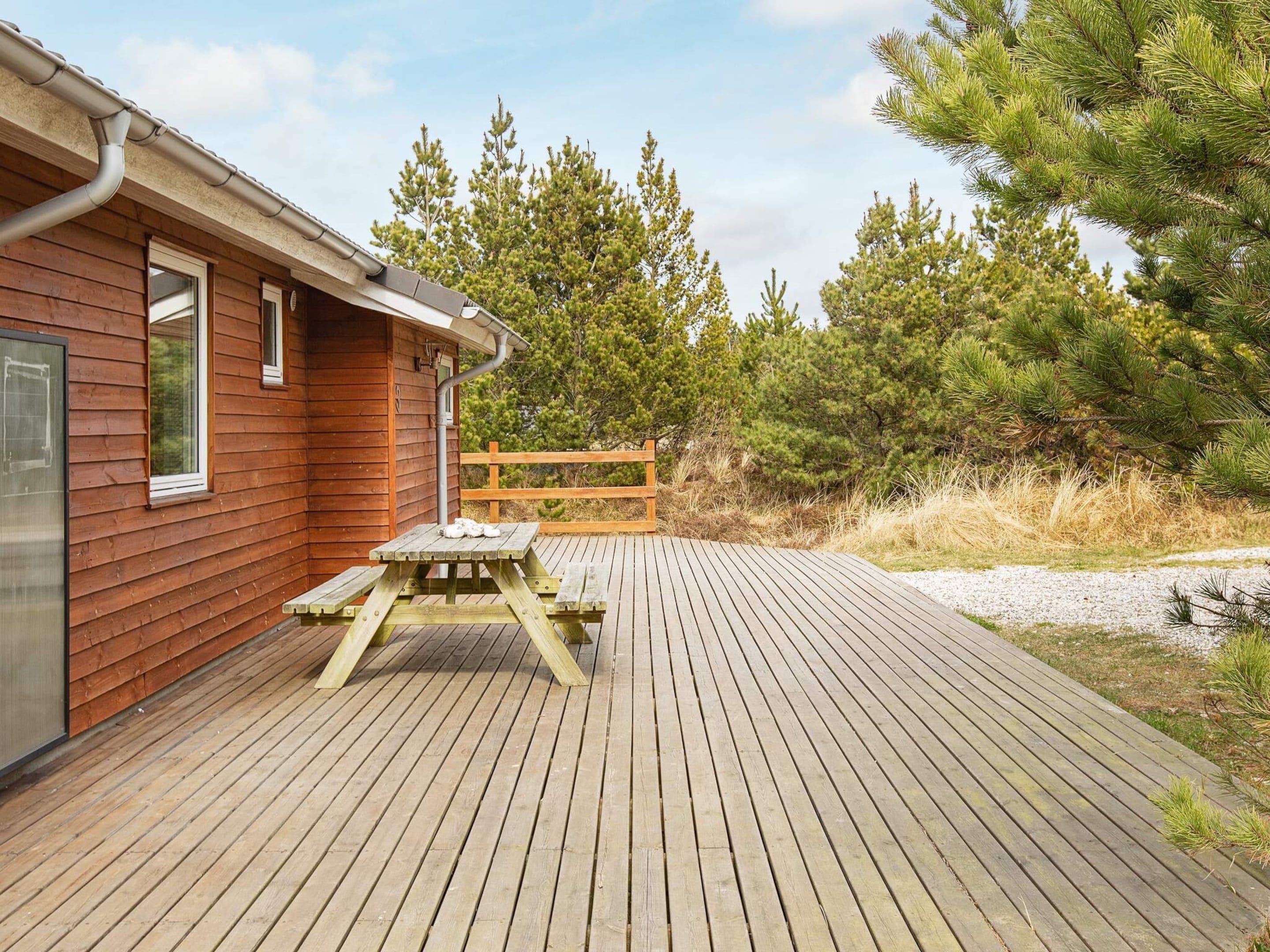 A wooden deck with a picnic table and benches, surrounded by trees and a building with windows.