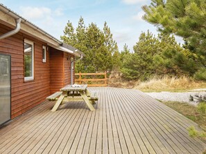 Una terraza de madera con una mesa de picnic y bancos, rodeada de árboles y un edificio con ventanas.