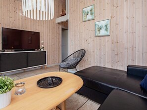 Una sala de estar moderna con un panel de pared de madera, un televisor de pantalla plana grande, un sofá de cuero negro, una silla de mimbre y una mesa de comedor de madera con un control remoto.