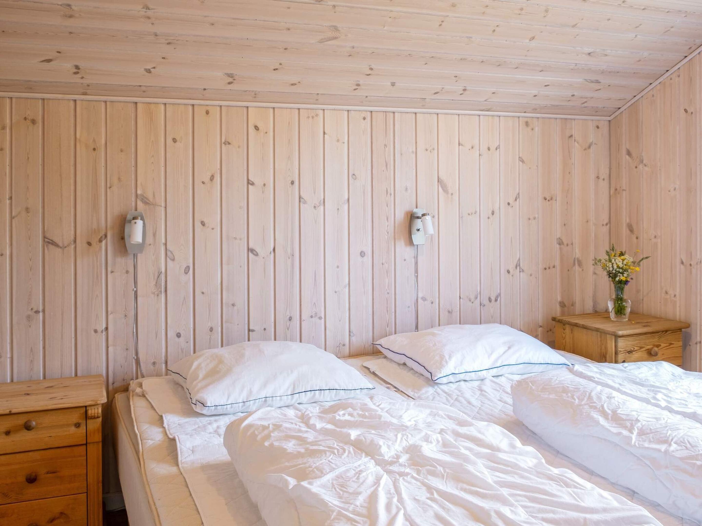 Une pièce aux murs lambrés, un lit recouvert de linge de maison blanc, une table de chevet en bois ornée d’un vase de fleurs et deux appliques murales.