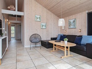A modern living room with a wooden ceiling, a black sofa with blue cushions, a round wooden coffee table, and a black wire chair.