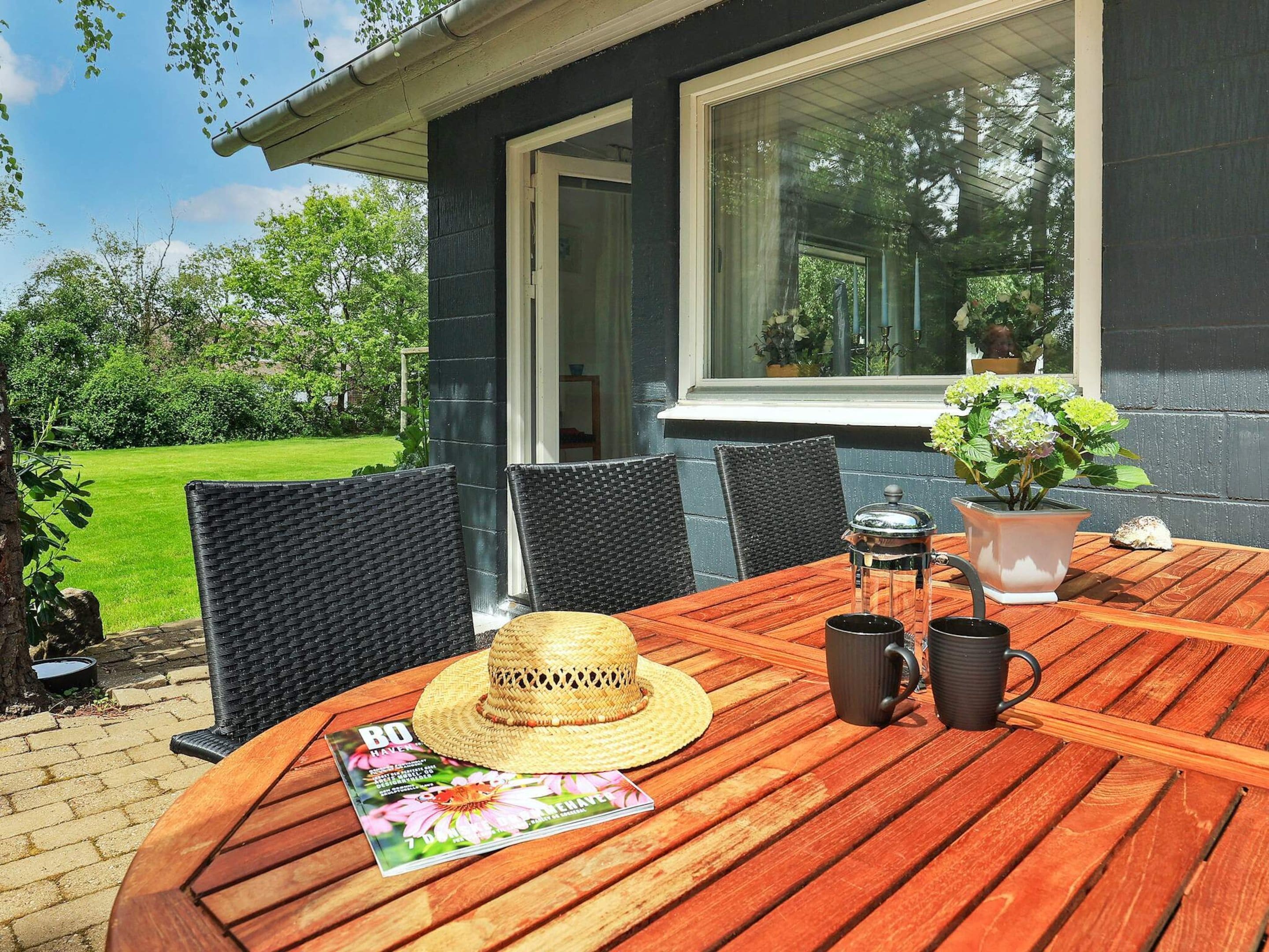 A wooden table with chairs, a magazine, a straw hat, and a coffee pot on a patio with a view of a garden and a building.