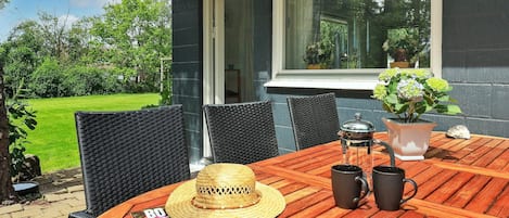 A wooden table with chairs, a magazine, a straw hat, and a coffee pot on a patio with a view of a garden and a building.