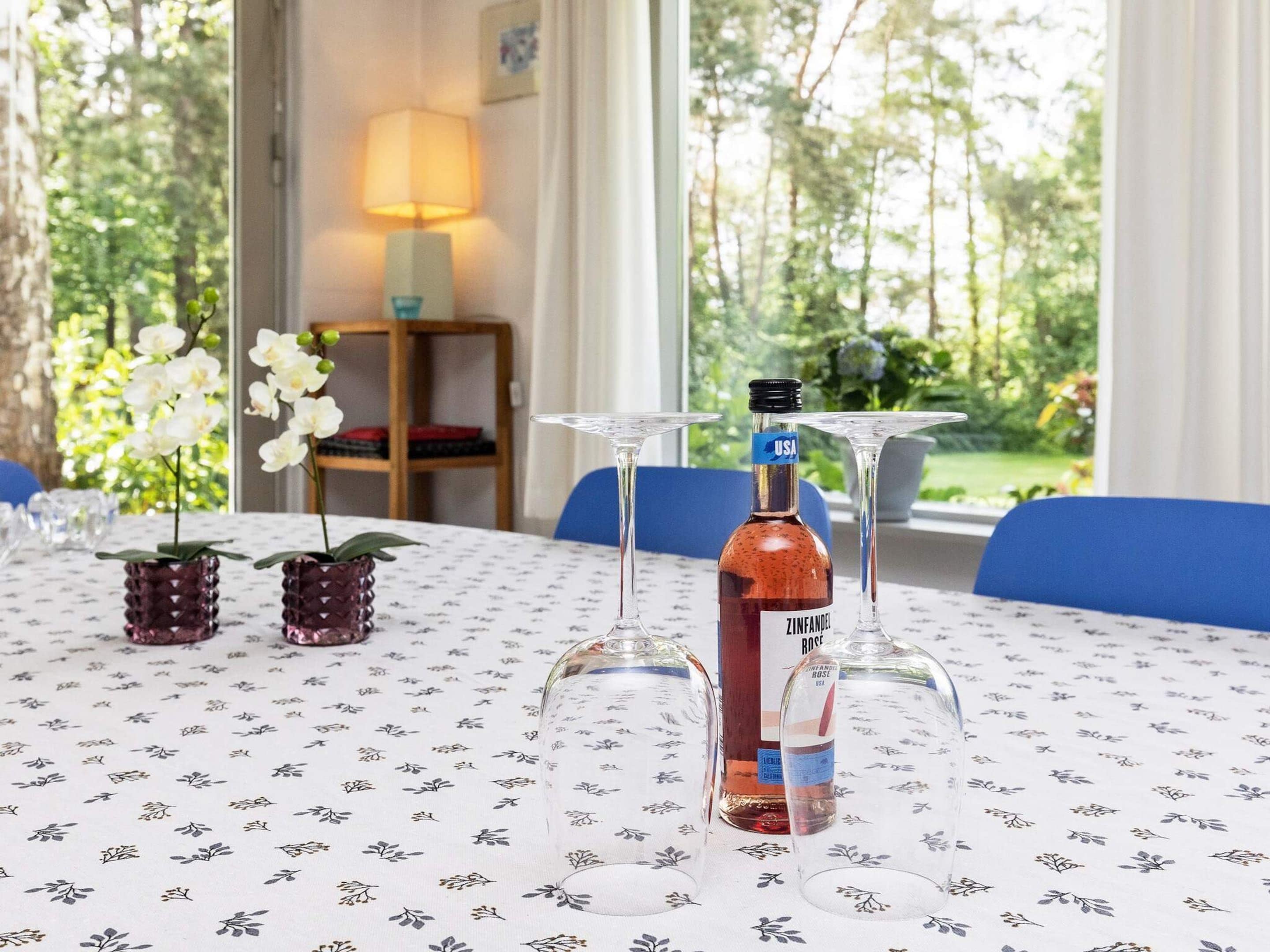 A table with a bottle of Zinfandel Rose wine, two empty wine glasses, and two orchid plants in decorative pots.