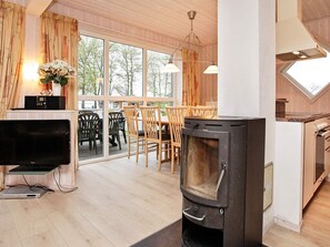 A cozy living room with a fireplace, a dining area with wooden furniture, and a large window offering a view of trees. - 8 Person Holiday Home in Gelting (Gelting)