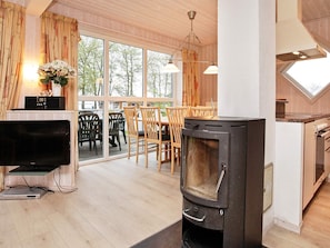 A cozy living room with a fireplace, a dining area with wooden furniture, and a large window offering a view of trees. - 8 Person Holiday Home in Gelting (Gelting)