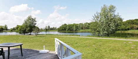 A wooden deck with a table and chairs, a white railing, and a view of a lake surrounded by trees.