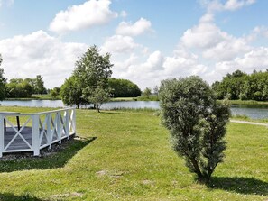 A park with a wooden deck, a small tree, and a lake. - Holiday Home in Otterndorf (Otterndorf)