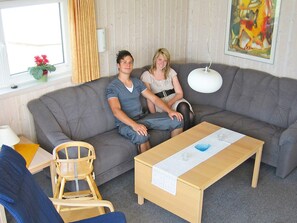 Una habitación con un sofá gris, una mesa de centro de madera con un posavasos, una silla de madera y una ventana con cortinas.