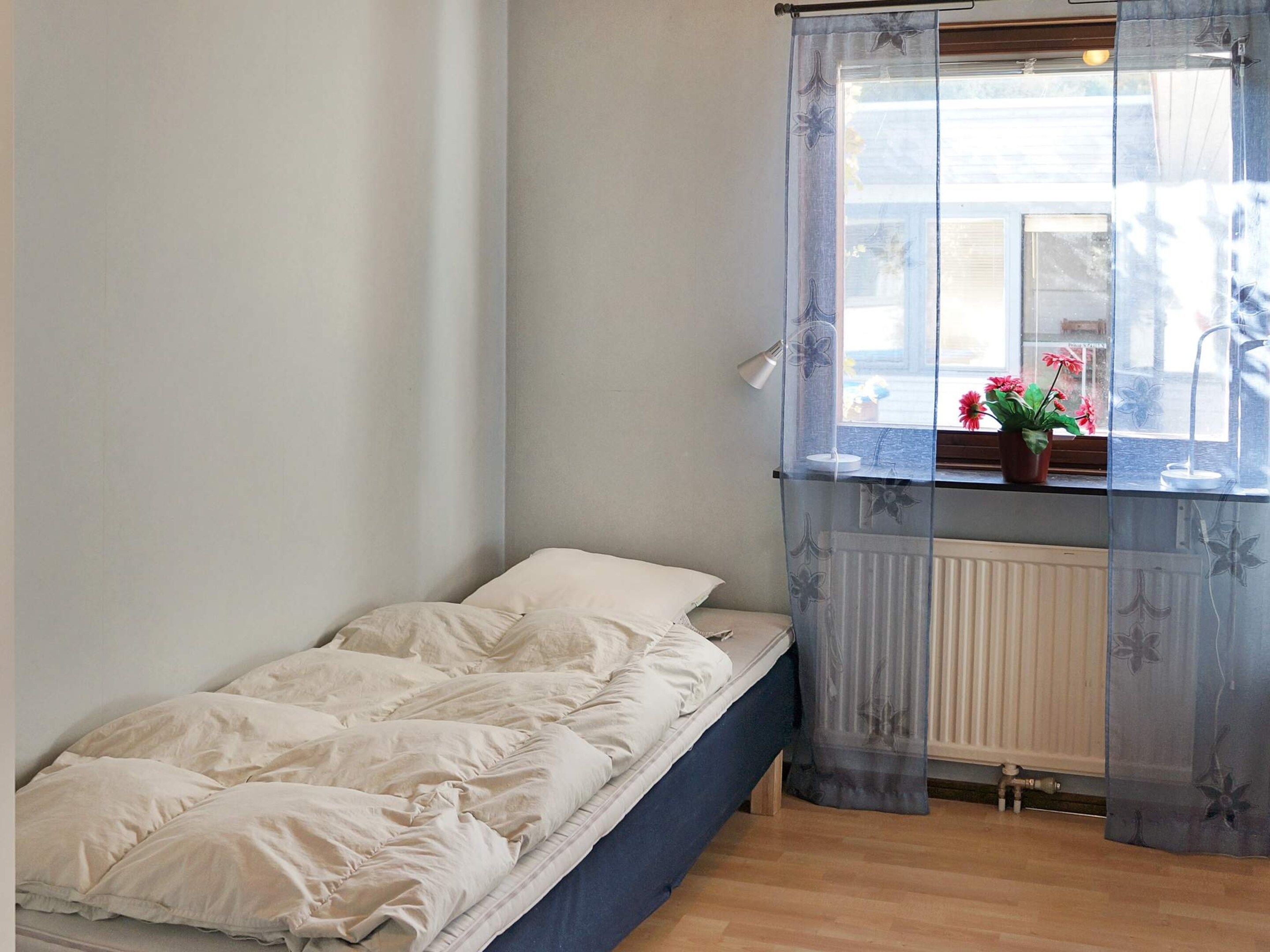 Yhden hengen sänky, jossa on valkoinen peitto ja sininen pääty, huoneessa, jonka ikkuna on peitetty kukkakuviollisella, läpikuultavalla verholla.