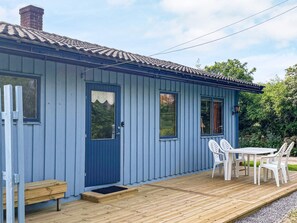 A small, single-story building with a blue exterior, a front door, a window, and a deck area with a table and chairs.