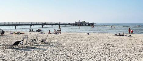 A beach with a pier extending into the water, sun loungers, and people enjoying the seaside.