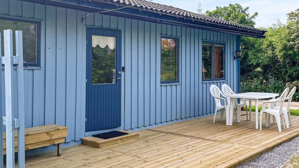 A small, single-story building with a blue exterior, a front door with a window, a small porch with a table and chairs, and a wooden bench. - 4 Star Holiday Home in Kopingsvik (Kopingsvik)