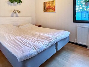 A neatly made bed with a white duvet and pillows, a wooden headboard, a painting on the wall, and a window with a view of a house. - 4 Star Holiday Home in Kopingsvik (Kopingsvik)