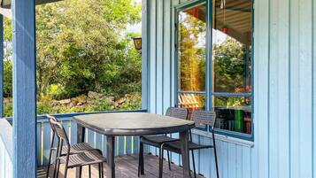 A covered outdoor seating area with a table and chairs, a view of trees and a garden, and a building with blue siding.