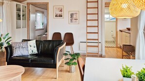 A modern living room with a black leather sofa, wooden coffee table, and a dining area with a white table and chairs. There are potted plants and a ladder in the room. - 6 Person Holiday Home in Gudhjem (Gudhjem)