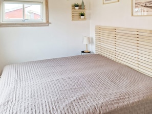 A bedroom with a bed, a window, a bedside table with a lamp, and a wall-mounted shelf with plants.
