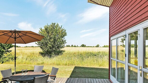A wooden deck with a table and chairs, an umbrella, and a view of a field and tree. - 6 Person Holiday Home in Gudhjem (Gudhjem)