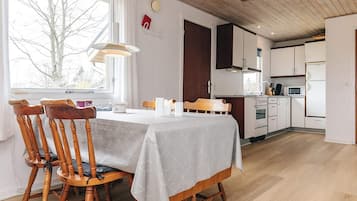 A dining area with a table set for two, wooden chairs, a window with a view of trees, and a kitchen with white cabinets and appliances.
