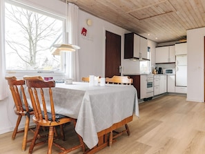 A dining area with a table set for two, wooden chairs, a window with a view of trees, and a kitchen with white cabinets and appliances.