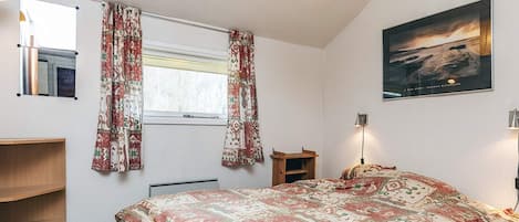 A bedroom with a bed, a window with curtains, a wooden shelf, a bedside table, a lamp, and a framed picture on the wall.