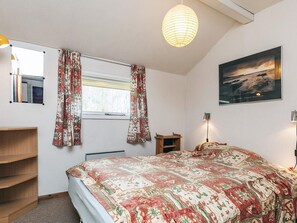 A bedroom with a bed, a window with curtains, a wooden shelf, a bedside table, a lamp, and a framed picture on the wall.