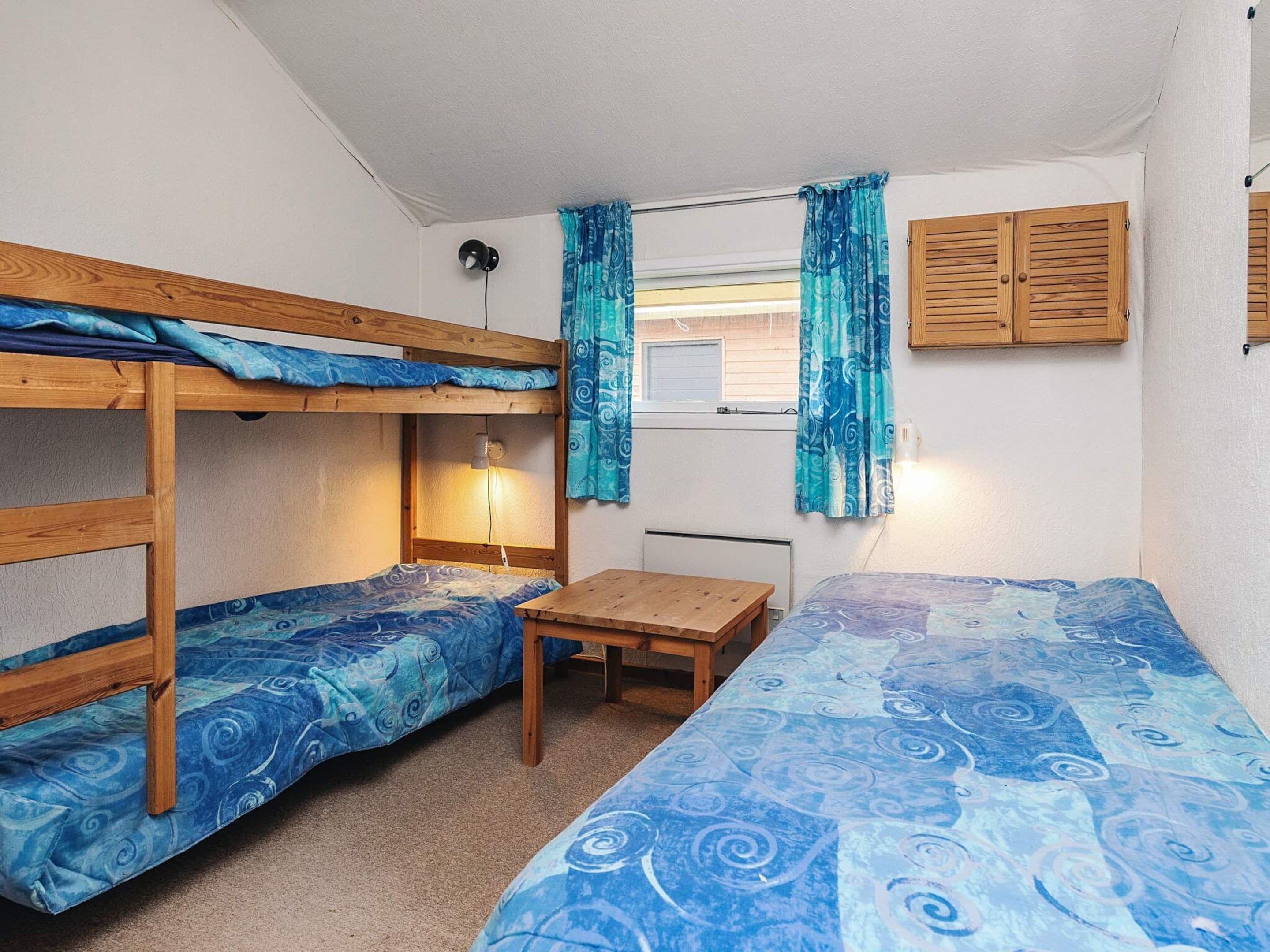 A dormitory room with bunk beds, a single bed, a small table, and a window with curtains.