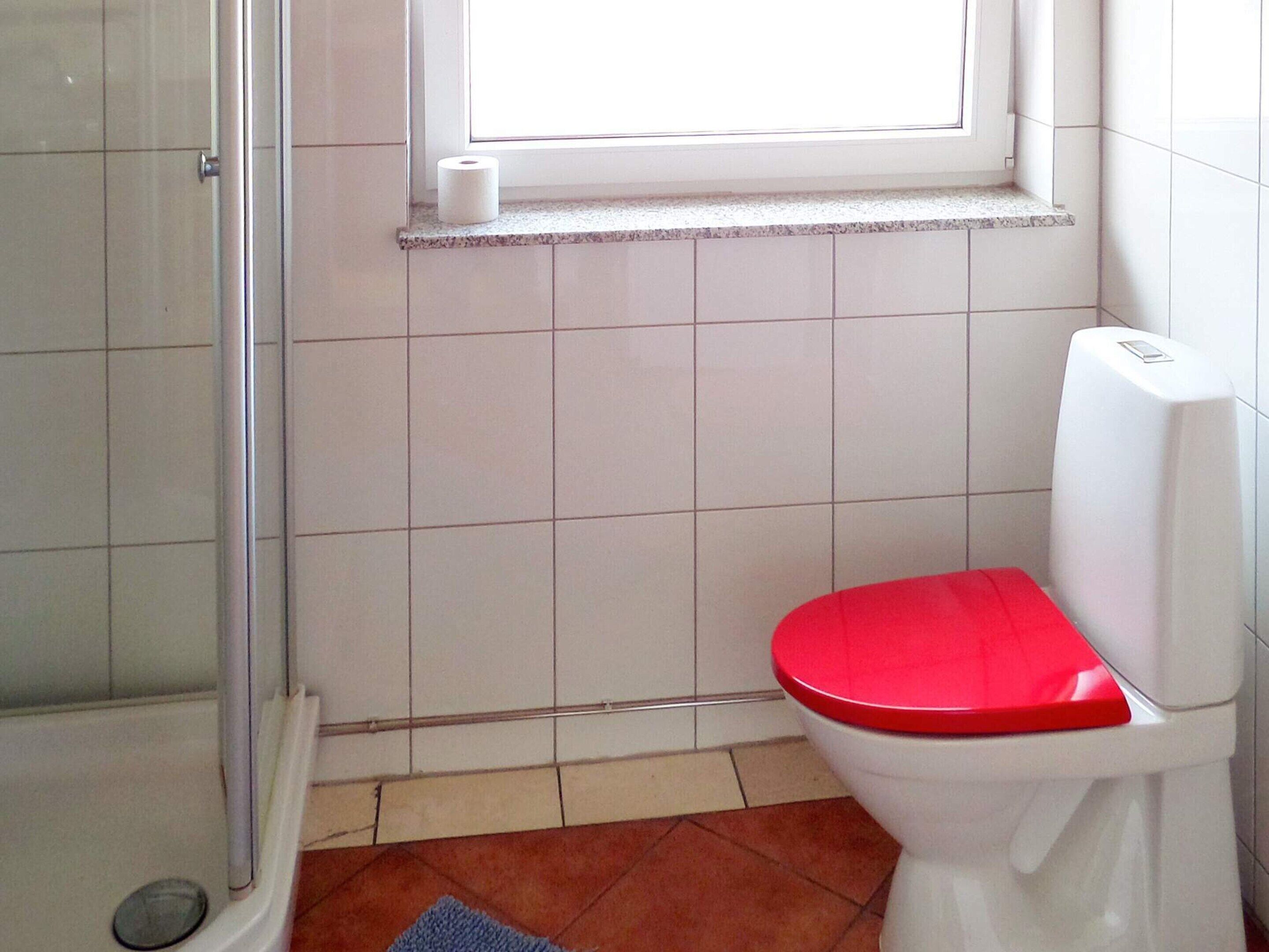 A bathroom with a white toilet, a red seat, a shower area, and a window above the toilet.