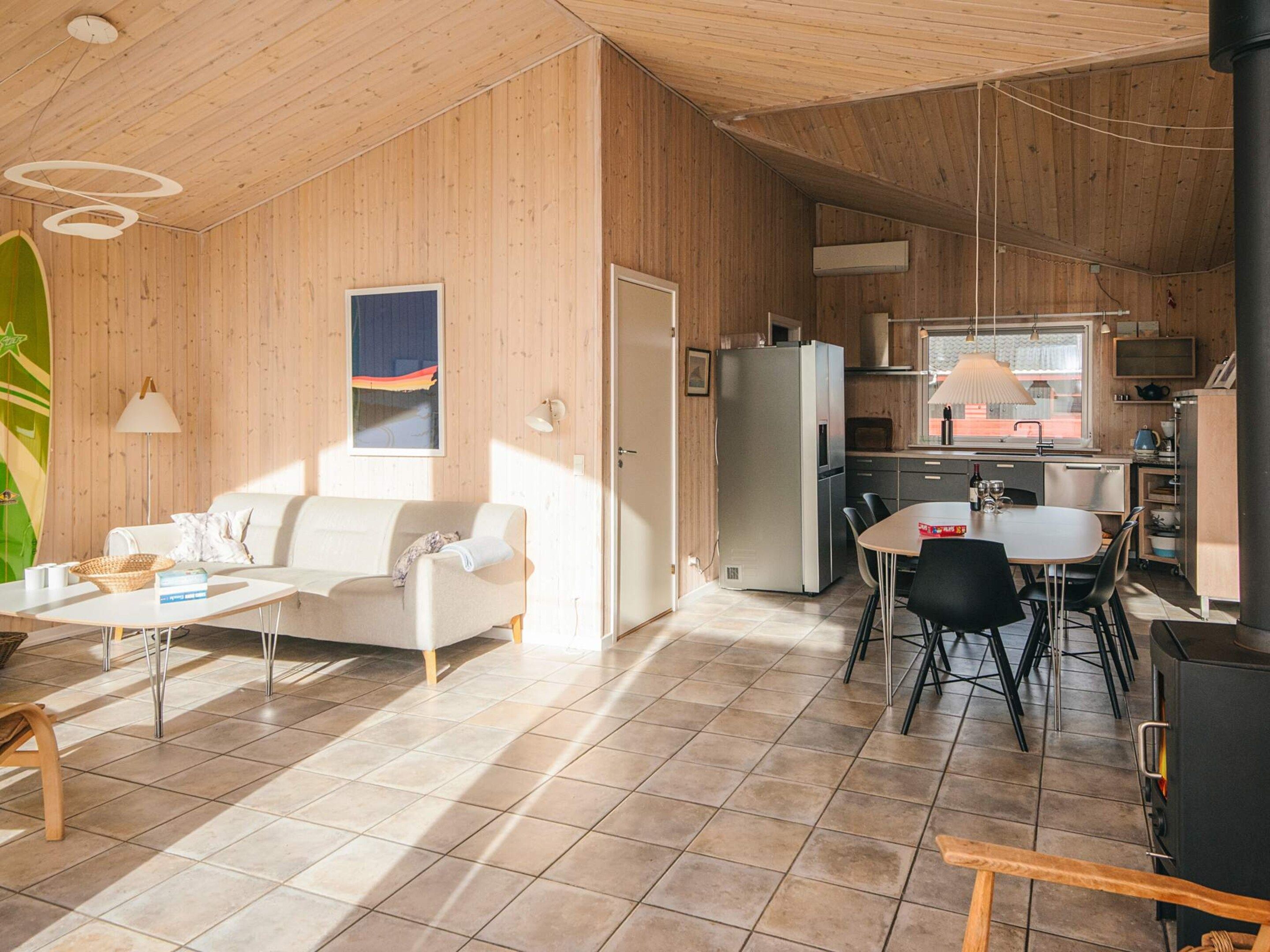 A modern interior with a wooden ceiling, a dining area with a table and chairs, a kitchen with a refrigerator and stove, and a sofa in the living area.