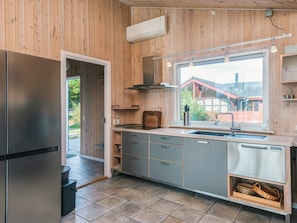 Uma cozinha moderna com paredes de madeira, uma geladeira de aço inoxidável, armários cinzas, lava-louças embutida e uma janela com vista para uma casa e vegetação.