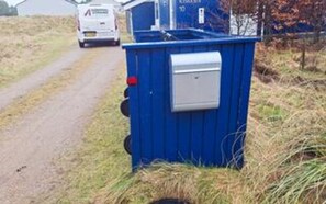 Een blauwe mobiele toilet met een wit deksel, vlakbij een weg met een wit voertuig op de achtergrond.