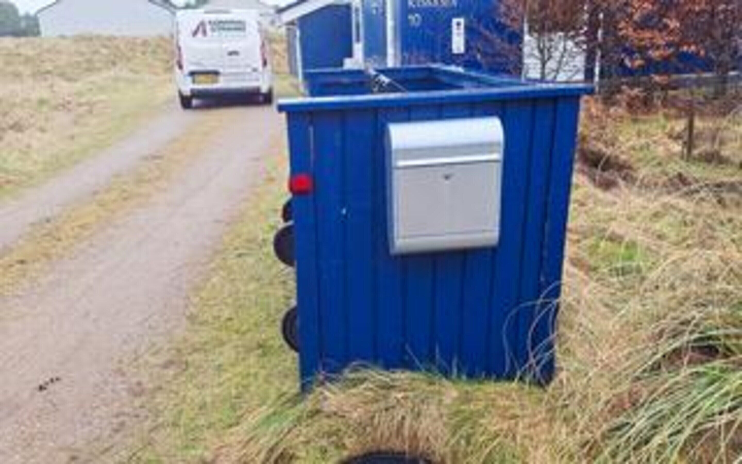 A blue portable toilet with a white lid, situated beside a road with a white vehicle in the background.