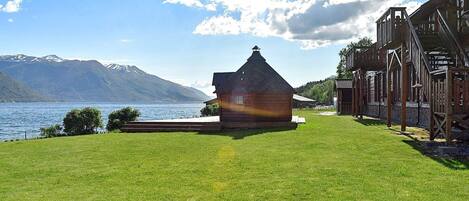 A wooden cabin with a conical roof by a lake, surrounded by a well-maintained lawn and a wooden structure with stairs.