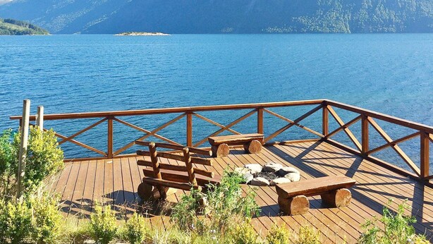 Une terrasse en bois, dotée de bancs et d’un foyer, offre une vue sur un lac, avec des montagnes en toile de fond.