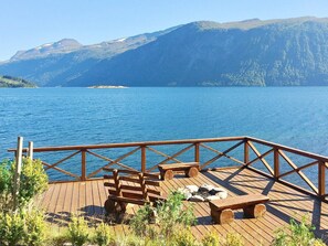 Eine Holzterrasse mit Bänken und einem Feuerkorb mit Blick auf einen See, im Hintergrund Berge.