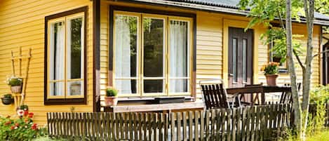 A yellow wooden house with large windows, a wooden porch, and a picket fence.