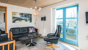 A modern living room with a black leather sofa, a round table, and a black office chair. There is a large window with a view of trees and buildings outside. - 12 Person Holiday Home in Hojslev-by Traum (Hojslev)