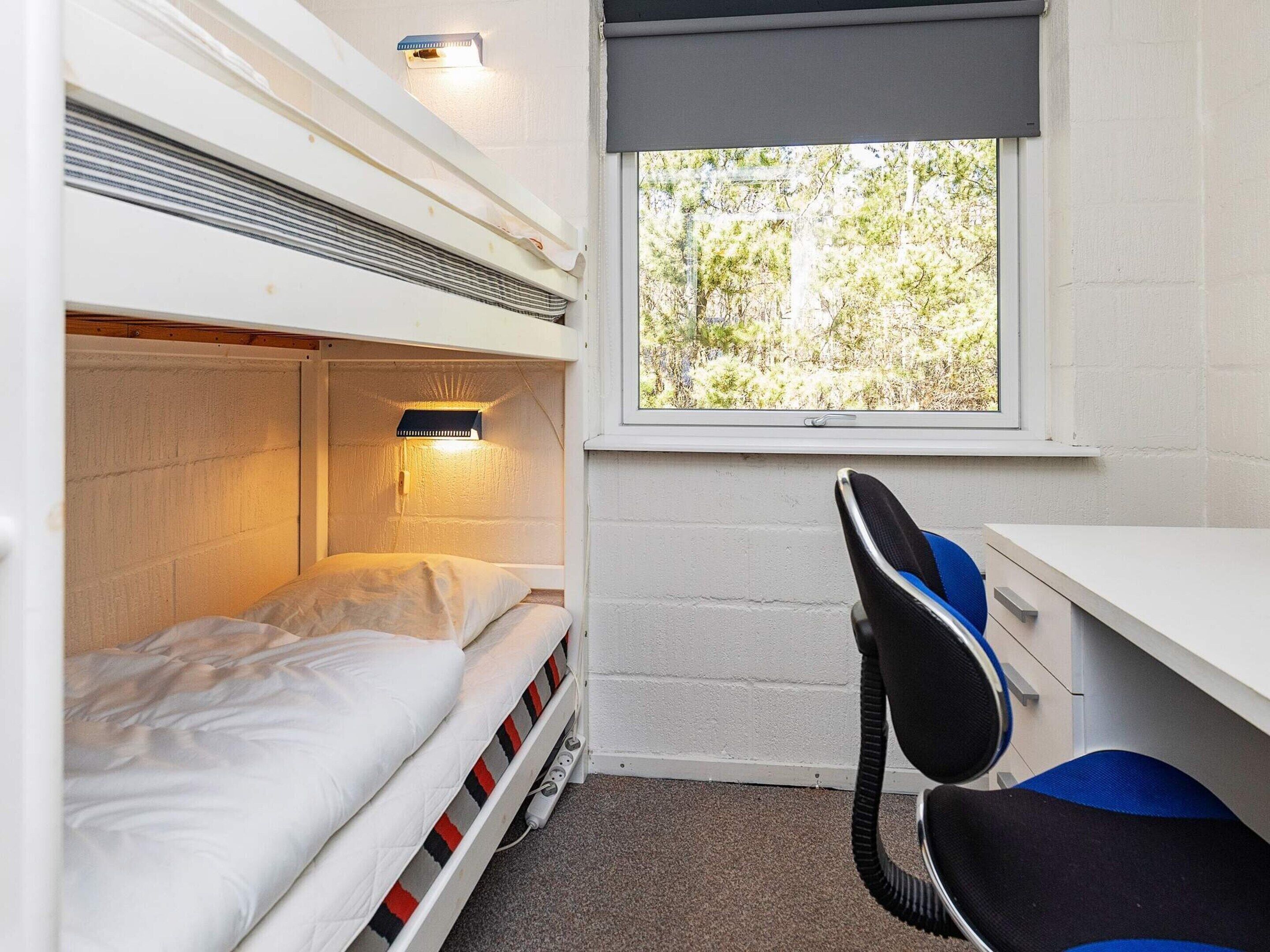 A bunk bed with a desk and chair, a window with a view of trees, and a wall-mounted heater.