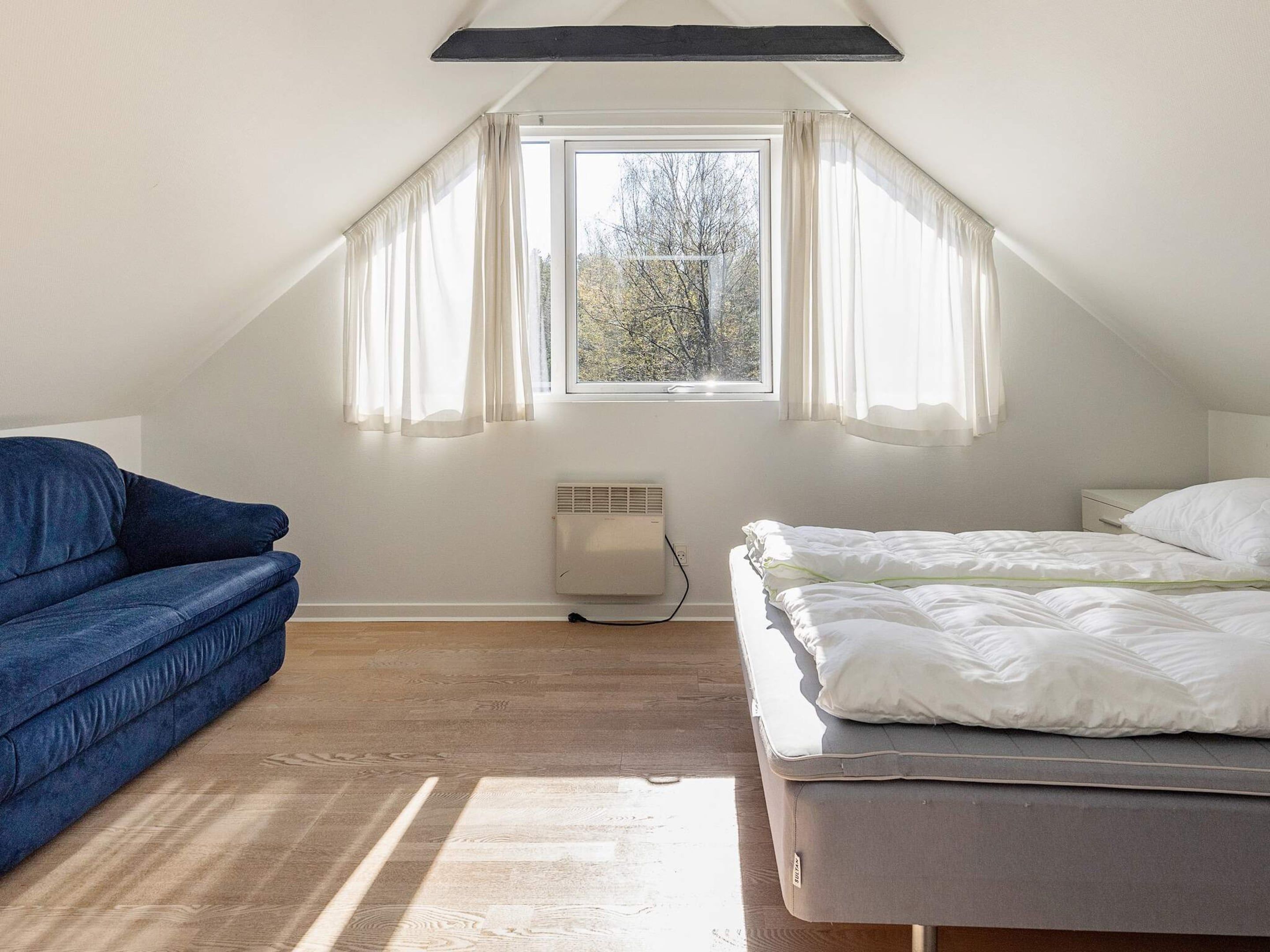 A room with a sloped ceiling, a bed with white bedding, a blue sofa, and a window with curtains.