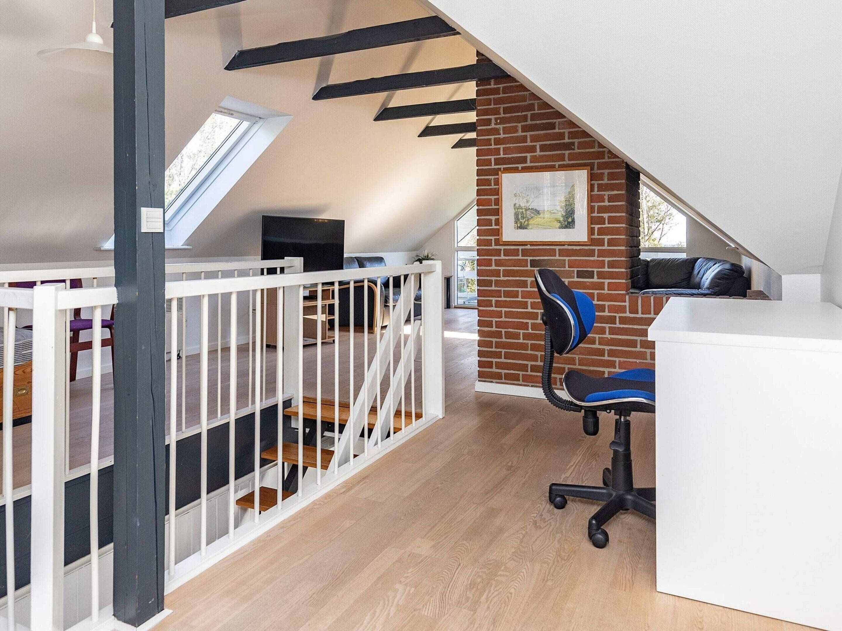 A loft bedroom with a sloped ceiling, a brick wall, a white desk, a blue office chair, and a white crib.