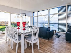 A dining area with a glass table and white chairs, large windows with a view, and a dark blue sofa. - 7 Person Holiday Home in Stege (Stege)